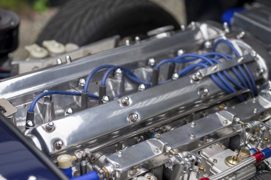 A close-up of a polished metal engine with visible blue ignition wires and various metallic components. The engine features prominent bolts and a sleek, reflective surface with detailed mechanical parts.