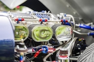 Close-up view of a car engine compartment showing multiple shiny chrome air filters labeled with a logo, connected with red and blue hoses and various metallic components. The focus is on the intricate and reflective engine parts under bright lighting.