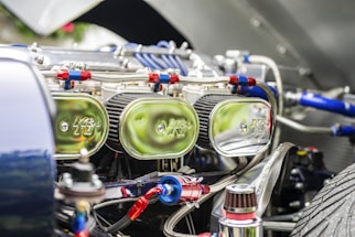 Close-up view of a car engine compartment showing multiple shiny chrome air filters labeled with a logo, connected with red and blue hoses and various metallic components. The focus is on the intricate and reflective engine parts under bright lighting.