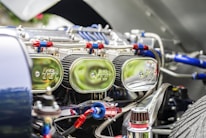 Close-up view of a car engine compartment showing multiple shiny chrome air filters labeled with a logo, connected with red and blue hoses and various metallic components. The focus is on the intricate and reflective engine parts under bright lighting.