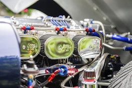 Close-up view of a car engine compartment showing multiple shiny chrome air filters labeled with a logo, connected with red and blue hoses and various metallic components. The focus is on the intricate and reflective engine parts under bright lighting.