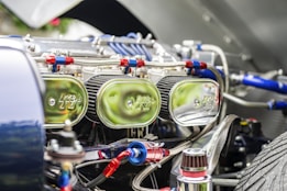 Close-up view of a car engine compartment showing multiple shiny chrome air filters labeled with a logo, connected with red and blue hoses and various metallic components. The focus is on the intricate and reflective engine parts under bright lighting.