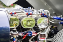 Close-up view of a car engine compartment showing multiple shiny chrome air filters labeled with a logo, connected with red and blue hoses and various metallic components. The focus is on the intricate and reflective engine parts under bright lighting.