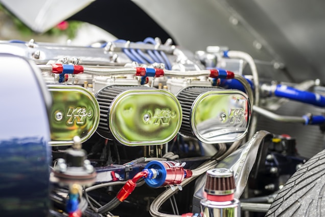 Close-up view of a car engine compartment showing multiple shiny chrome air filters labeled with a logo, connected with red and blue hoses and various metallic components. The focus is on the intricate and reflective engine parts under bright lighting.