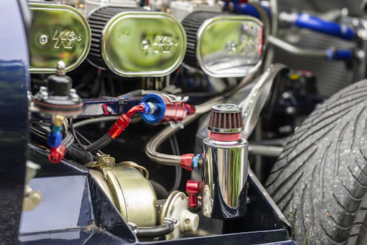 Close-up view of a vehicle engine with several mechanical components, including a polished metal canister and visible air filters. The engine is intricately detailed with multiple hoses, connections, and a prominent, large treaded tire visible in the background.