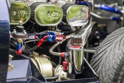 Close-up view of a vehicle engine with several mechanical components, including a polished metal canister and visible air filters. The engine is intricately detailed with multiple hoses, connections, and a prominent, large treaded tire visible in the background.