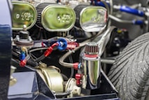 Close-up view of a vehicle engine with several mechanical components, including a polished metal canister and visible air filters. The engine is intricately detailed with multiple hoses, connections, and a prominent, large treaded tire visible in the background.