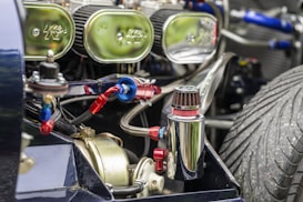 Close-up view of a vehicle engine with several mechanical components, including a polished metal canister and visible air filters. The engine is intricately detailed with multiple hoses, connections, and a prominent, large treaded tire visible in the background.