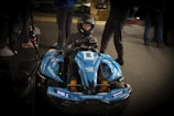 A person wearing a helmet is sitting in a blue go-kart labeled with the number 13. The go-kart has multiple stickers and decals, including '#BAKKERUDBLUE' and 'informativ'. The scene appears to be indoors, with several other people standing around in the background.