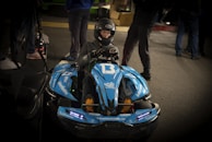 A person wearing a helmet is sitting in a blue go-kart labeled with the number 13. The go-kart has multiple stickers and decals, including '#BAKKERUDBLUE' and 'informativ'. The scene appears to be indoors, with several other people standing around in the background.