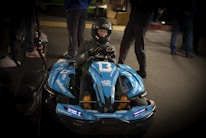 A person wearing a helmet is sitting in a blue go-kart labeled with the number 13. The go-kart has multiple stickers and decals, including '#BAKKERUDBLUE' and 'informativ'. The scene appears to be indoors, with several other people standing around in the background.