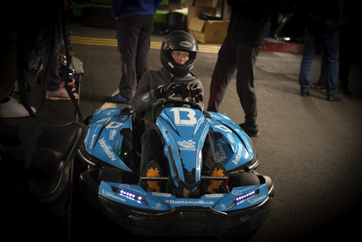 A person wearing a helmet is sitting in a blue go-kart labeled with the number 13. The go-kart has multiple stickers and decals, including '#BAKKERUDBLUE' and 'informativ'. The scene appears to be indoors, with several other people standing around in the background.