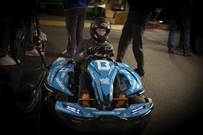 A person wearing a helmet is sitting in a blue go-kart labeled with the number 13. The go-kart has multiple stickers and decals, including '#BAKKERUDBLUE' and 'informativ'. The scene appears to be indoors, with several other people standing around in the background.