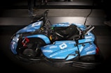 A sleek blue go-kart with various sponsorship logos is parked indoors on a dark surface. The go-kart features a single seat, a steering wheel, and a visible structure designed for performance and speed.