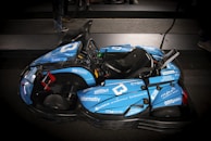 A sleek blue go-kart with various sponsorship logos is parked indoors on a dark surface. The go-kart features a single seat, a steering wheel, and a visible structure designed for performance and speed.