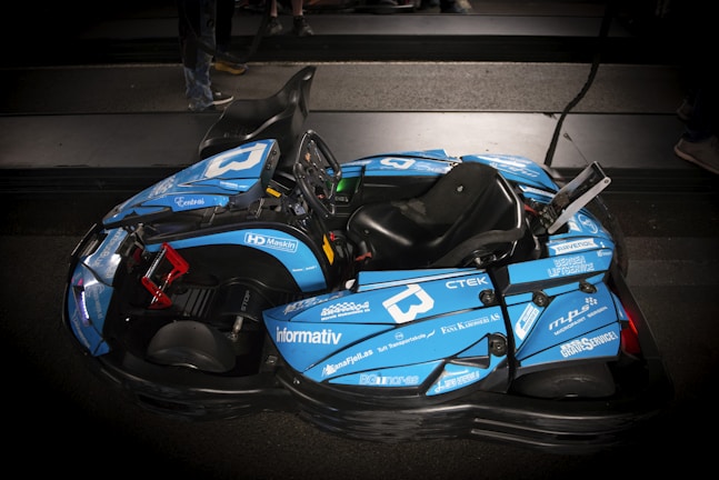 A sleek blue go-kart with various sponsorship logos is parked indoors on a dark surface. The go-kart features a single seat, a steering wheel, and a visible structure designed for performance and speed.