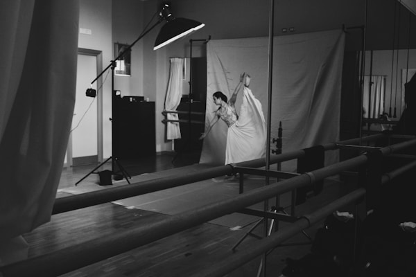 A dancer is performing a graceful pose, balancing on one foot in a studio setting. The room is equipped with professional photography lighting and a large backdrop. There are mirrors on one side of the room and ballet barres along the walls.