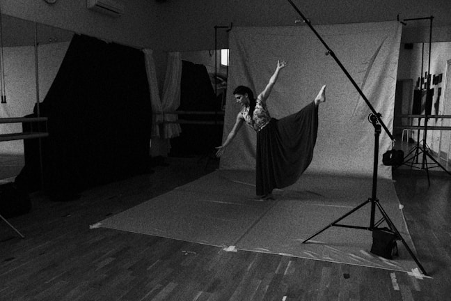 A dancer performs an elegant pose in a studio setting, captured in black and white. The room contains a backdrop, studio lights, and reflective surfaces, with wooden floors and various photography equipment visible. The dancer is balanced gracefully, one leg extended, wearing a flowing skirt.