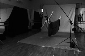 A dancer performs an elegant pose in a studio setting, captured in black and white. The room contains a backdrop, studio lights, and reflective surfaces, with wooden floors and various photography equipment visible. The dancer is balanced gracefully, one leg extended, wearing a flowing skirt.