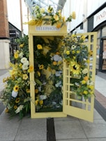 A traditional telephone booth is creatively adorned with an array of flowers in various shades of yellow, white, and green. The floral arrangement includes sunflowers, roses, and other botanical elements, giving the booth a vibrant and lively appearance. The booth is set in an outdoor shopping area, surrounded by brick buildings and store signage.