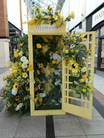 A traditional telephone booth is creatively adorned with an array of flowers in various shades of yellow, white, and green. The floral arrangement includes sunflowers, roses, and other botanical elements, giving the booth a vibrant and lively appearance. The booth is set in an outdoor shopping area, surrounded by brick buildings and store signage.