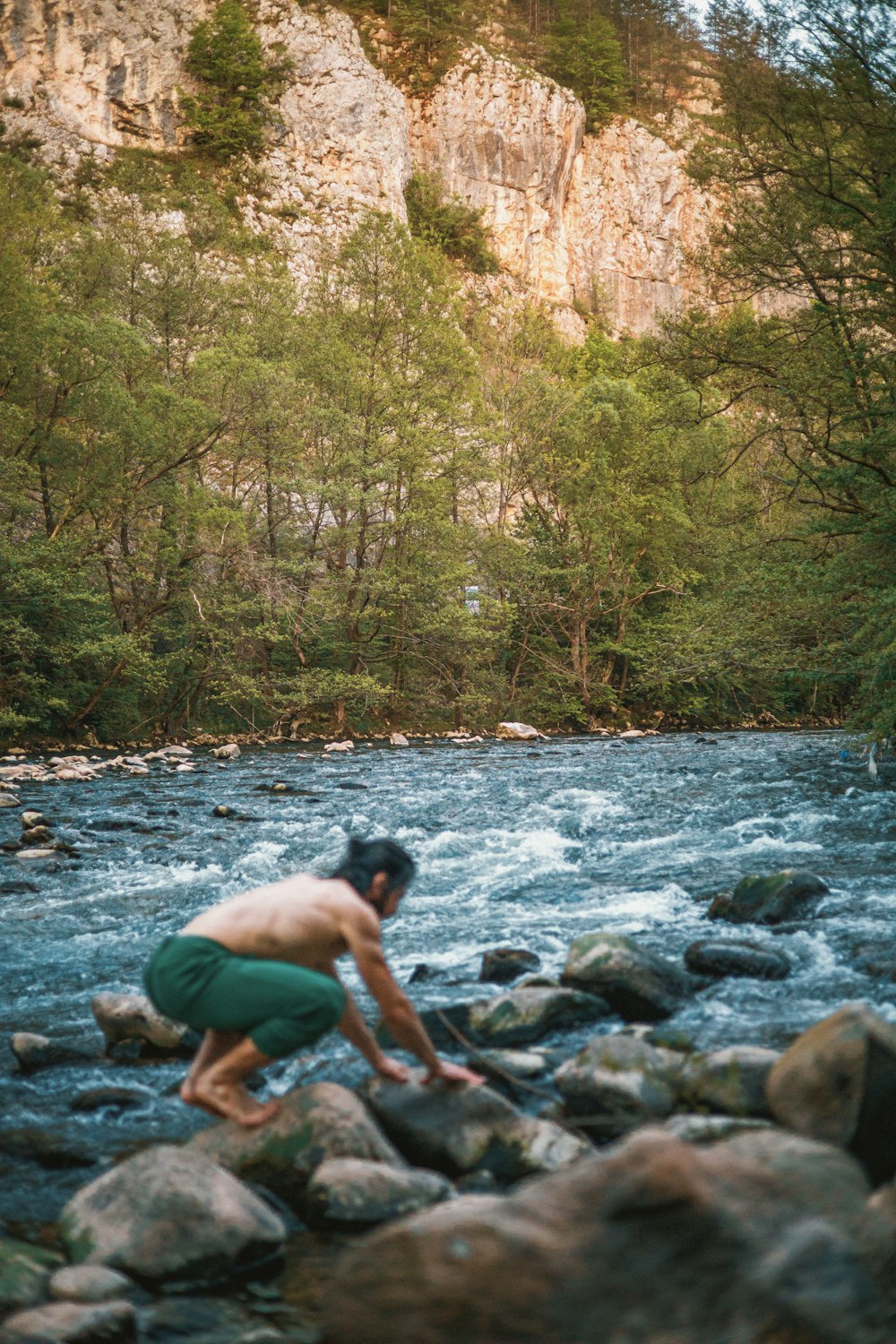 a shirtless man crouches on rocks near a river