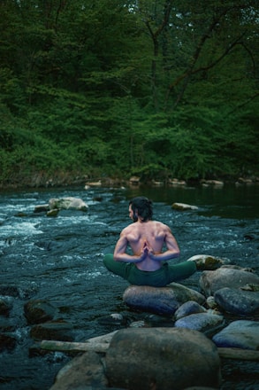 A person sits on a rock in the middle of a flowing stream, surrounded by lush greenery. The individual is in a yoga pose with hands joined behind their back, showcasing a serene and meditative posture. The environment is tranquil, with the soft movement of water and the dense forest creating a peaceful ambiance.