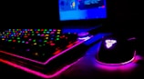 A dimly lit gaming setup showcasing a colorful RGB keyboard and a mouse with glowing accents. The background features a partially visible laptop screen.