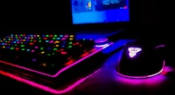 A dimly lit gaming setup showcasing a colorful RGB keyboard and a mouse with glowing accents. The background features a partially visible laptop screen.
