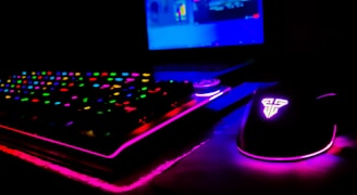 A dimly lit gaming setup showcasing a colorful RGB keyboard and a mouse with glowing accents. The background features a partially visible laptop screen.
