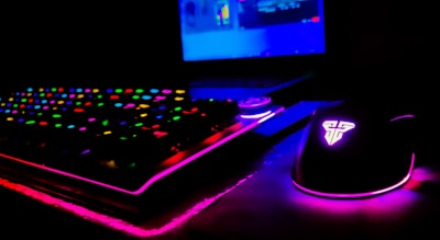 A dimly lit gaming setup showcasing a colorful RGB keyboard and a mouse with glowing accents. The background features a partially visible laptop screen.
