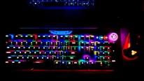 A brightly lit RGB gaming keyboard displays a spectrum of colors across its keys, creating a vibrant glow. A power button with a bright circular light is visible on the side, and a gaming mouse with similar RGB lighting sits adjacent to the keyboard. The top of a laptop is faintly visible above the keyboard.