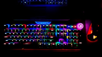 A brightly lit RGB gaming keyboard displays a spectrum of colors across its keys, creating a vibrant glow. A power button with a bright circular light is visible on the side, and a gaming mouse with similar RGB lighting sits adjacent to the keyboard. The top of a laptop is faintly visible above the keyboard.