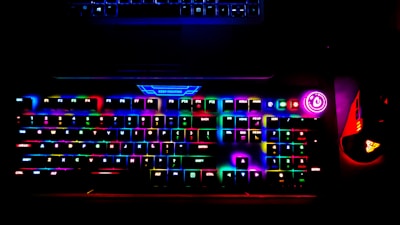 A brightly lit RGB gaming keyboard displays a spectrum of colors across its keys, creating a vibrant glow. A power button with a bright circular light is visible on the side, and a gaming mouse with similar RGB lighting sits adjacent to the keyboard. The top of a laptop is faintly visible above the keyboard.