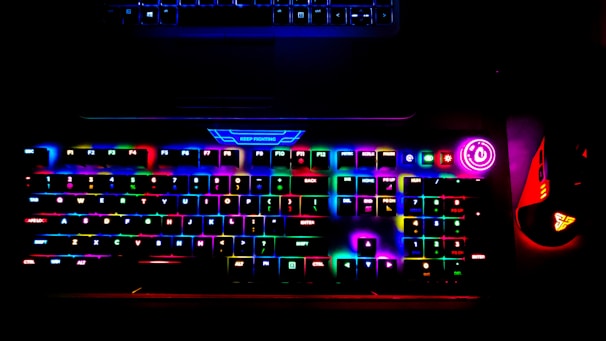 A brightly lit RGB gaming keyboard displays a spectrum of colors across its keys, creating a vibrant glow. A power button with a bright circular light is visible on the side, and a gaming mouse with similar RGB lighting sits adjacent to the keyboard. The top of a laptop is faintly visible above the keyboard.