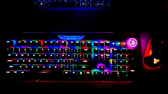 A brightly lit RGB gaming keyboard displays a spectrum of colors across its keys, creating a vibrant glow. A power button with a bright circular light is visible on the side, and a gaming mouse with similar RGB lighting sits adjacent to the keyboard. The top of a laptop is faintly visible above the keyboard.