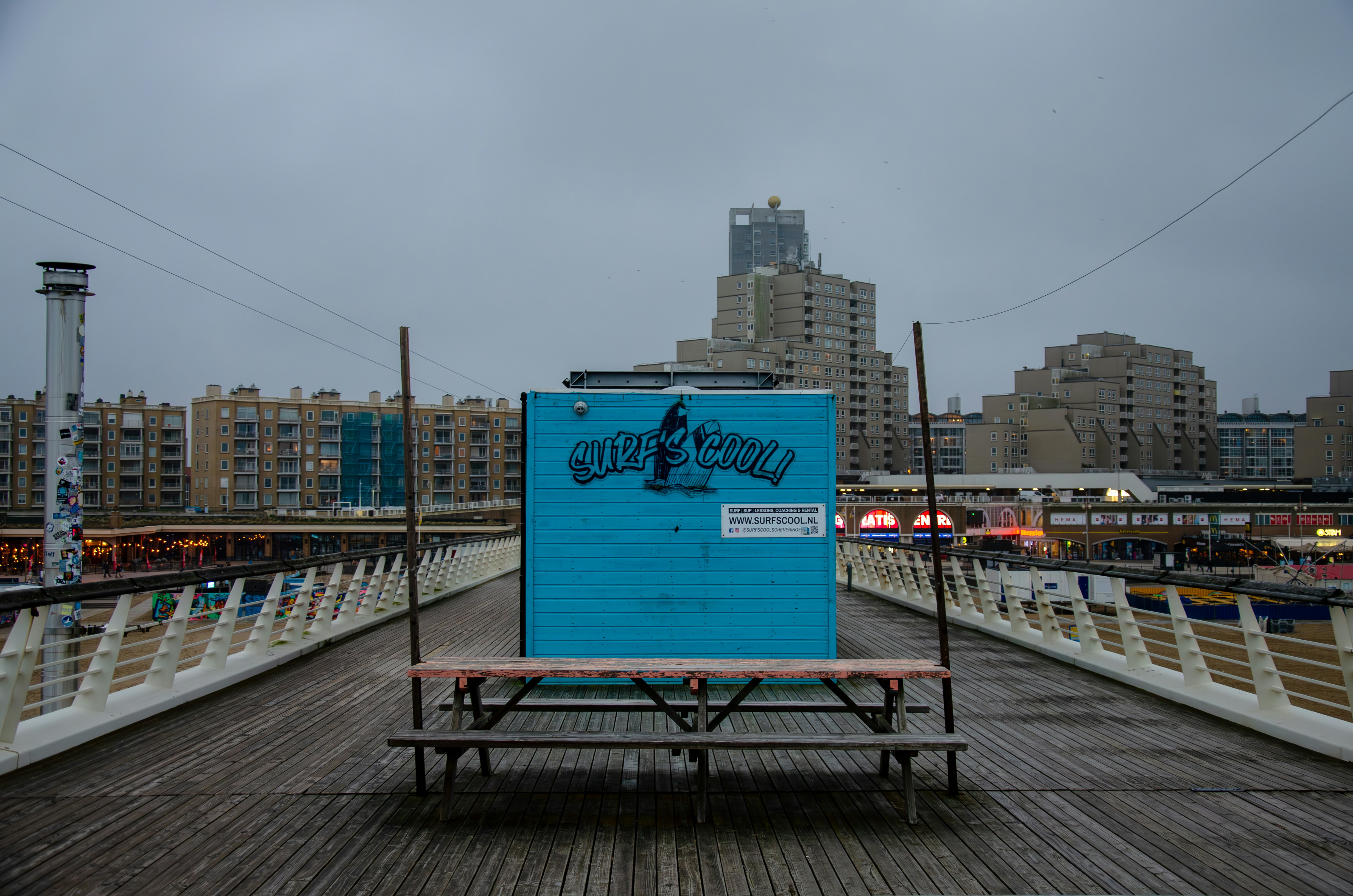 A blue wooden kiosk with graffiti-style text 'SURFSCOOL!' is positioned on a boardwalk. It stands in front of a row of modern, high-rise buildings under a cloudy sky. The kiosk is flanked by a simple bench and there are commercial signs in the background.