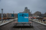A blue wooden kiosk with graffiti-style text 'SURFSCOOL!' is positioned on a boardwalk. It stands in front of a row of modern, high-rise buildings under a cloudy sky. The kiosk is flanked by a simple bench and there are commercial signs in the background.
