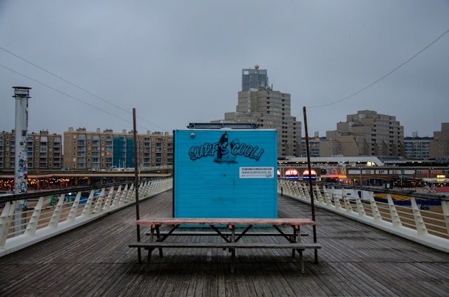 A blue wooden kiosk with graffiti-style text 'SURFSCOOL!' is positioned on a boardwalk. It stands in front of a row of modern, high-rise buildings under a cloudy sky. The kiosk is flanked by a simple bench and there are commercial signs in the background.