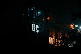 A dimly lit building is partially obscured by shadows and foliage. The letters 'UC' are prominently illuminated on the side of the structure. Warm lights can be seen through the windows and around the rooftop, suggesting a lively or vibrant interior, contrasting with the darkness outside.