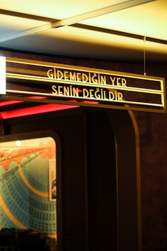 A vintage-looking sign with the text 'Gidemediğin Yer Senin Değildir' is illuminated in a yellowish glow. It is suspended from the ceiling with visible wires, and the background features parts of a colorful mural with blue and orange arcs intersecting and numbers visible.