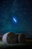A serene night sky filled with numerous stars, featuring a bright, vivid meteor streaking across the upper portion. Below, two large hay bales rest on an open field of grass, creating a contrast between the celestial and terrestrial elements.