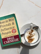 A copy of 'How to Win Friends & Influence People' by Dale Carnegie lies on a light-colored marble surface next to a cappuccino. The book cover is green and yellow with red accents, showing that it is a revised edition with over 15 million copies sold. The cappuccino is presented in a white cup on a saucer, featuring intricate latte art on top.