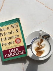 A copy of 'How to Win Friends & Influence People' by Dale Carnegie lies on a light-colored marble surface next to a cappuccino. The book cover is green and yellow with red accents, showing that it is a revised edition with over 15 million copies sold. The cappuccino is presented in a white cup on a saucer, featuring intricate latte art on top.