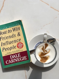 A copy of 'How to Win Friends & Influence People' by Dale Carnegie lies on a light-colored marble surface next to a cappuccino. The book cover is green and yellow with red accents, showing that it is a revised edition with over 15 million copies sold. The cappuccino is presented in a white cup on a saucer, featuring intricate latte art on top.