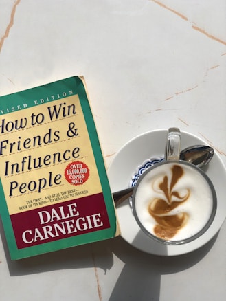 A copy of 'How to Win Friends & Influence People' by Dale Carnegie lies on a light-colored marble surface next to a cappuccino. The book cover is green and yellow with red accents, showing that it is a revised edition with over 15 million copies sold. The cappuccino is presented in a white cup on a saucer, featuring intricate latte art on top.