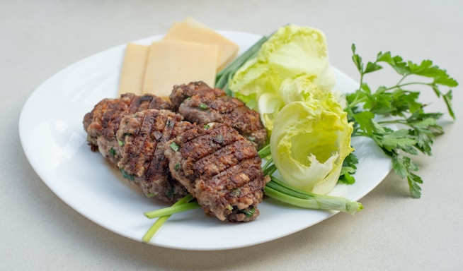 A white plate containing several grilled meat patties garnished with green onions, surrounded by slices of yellow cheese, lettuce leaves, and flat-leaf parsley.