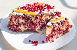 A cake on a white plate covered with powdered sugar and decorated with red currants, featuring a slice removed. The cake appears to be moist and layered, with visible fruit pieces throughout.