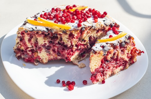 A cake on a white plate covered with powdered sugar and decorated with red currants, featuring a slice removed. The cake appears to be moist and layered, with visible fruit pieces throughout.