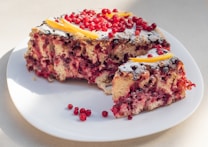 A cake featuring a dense, fruity interior with visible red berries, possibly cranberries or red currants, topped with a dusting of powdered sugar and garnished with thin lemon slices. It is situated on a white plate against a neutral background. A slice has been taken out, revealing the vibrant interior.